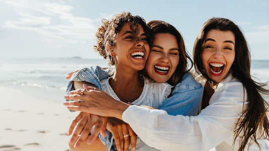 Three women laughing and embracing together on a sunny beach, with the ocean and bright blue sky in the background.