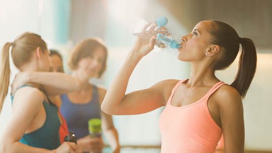 Woman drinking water after a workout while others rest in the background at a gym or fitness studio.