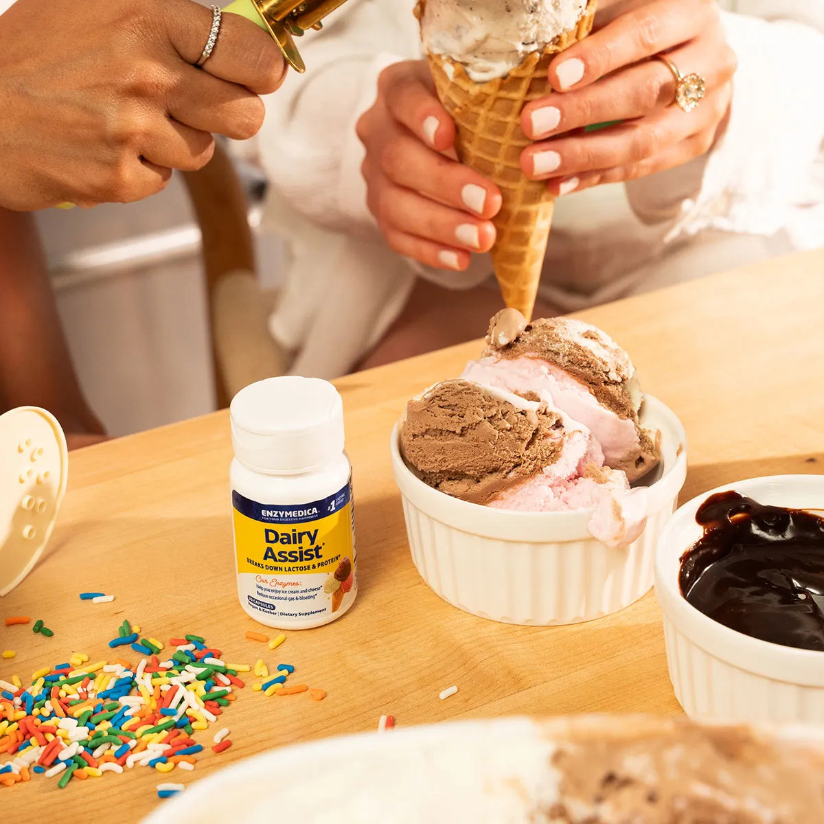 Bottle of Enzymedica Dairy Assist supplement on a table beside ice cream cones, sprinkles, and chocolate sauce.
