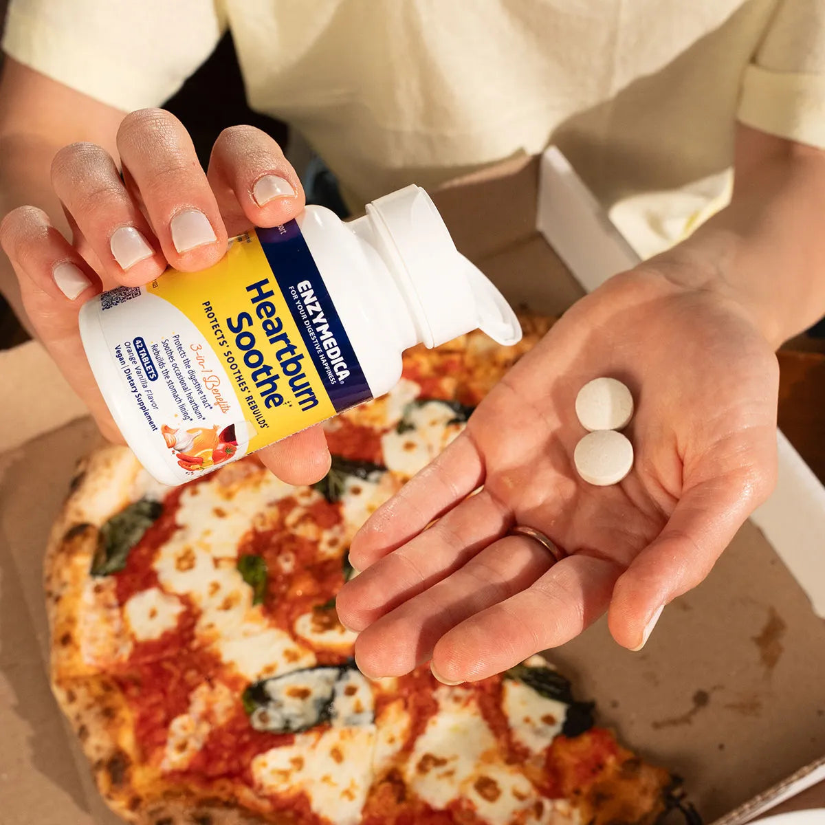 Person holding a bottle of Enzymedica Heartburn Soothe tablets and two chewable tablets above a pizza.